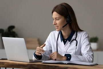 Telehealth. Confident female doctor attending physician in modern headset with mic sit by laptop in...
