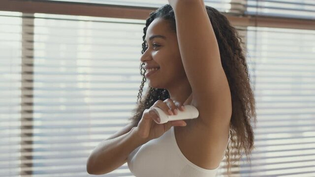 Personal hygiene concept. Young african american lady applying deodorant on armpit, smiling at bathroom, tracking shot