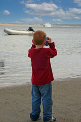 Young redhaired boy stood on beach looking at boat through binoculars