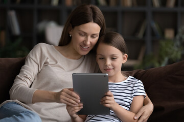 Using pad with mom. Focused preteen girl and young mommy nanny hug on couch look on touchpad screen engaged in easy distant learning. Friendly sisters adult elder and little kid watch video on tablet
