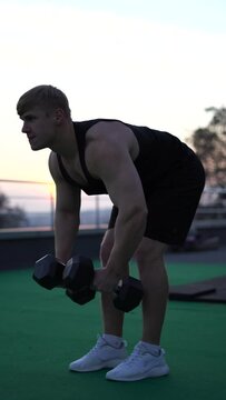 Caucasian white muscular man lifting dumbbells against the sunset sky background. Concept of willpower, motivation and passion. 4k vertical shot 