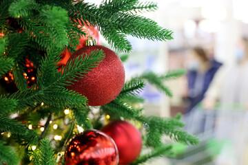 Christmas tree with red toy balls in a shopping mall on background of walking people in masks. New Year decorations, winter holidays during coronavirus pandemic