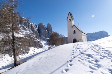 Fototapeta na wymiar Chiesetta innevata a Passo Gardena sulle Dolomiti in inverno, chiesetta stile alpino ai piedi delle vette del Gruppo Sella sulle Alpi Italiane