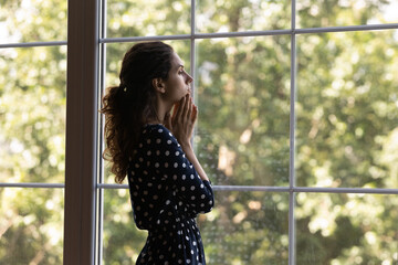 Frustrated unhappy anxious young hispanic woman looking in distance standing near huge window,...