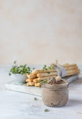 Homemade liver pate, spread or mousse in glass jar with grissini and microgreens, light concrete background.