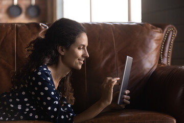 Side view smiling latin woman using digital touchpad, lying on cozy leather sofa, playing online games, web surfing information, choosing goods or services shopping in internet store, booking tickets.