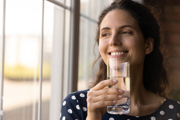 Dreamy peaceful young hispanic woman looking in distance out of window, drinking glass of fresh pure mineral water, enjoying morning daily healthcare lifestyle habit, staying refreshed during day. - obrazy, fototapety, plakaty