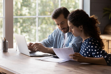 Concentrated young couple managing household budget or savings together, doing financial paperwork couple calculating domestic expenditures, paying utility bills using computer e-banking application.