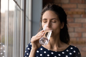 Peaceful young hispanic woman drinking glass of fresh pure water, preventing organism dehydration, keep dietary lifestyle, feeling thirsty standing near window at home, daily healthcare habit. - obrazy, fototapety, plakaty
