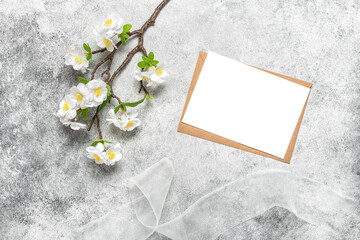 Spring stationery still life. Blank greeting card mockup, brown envelope and cherry blossom decorative branch. Gray grunge background. Top view, flat lay.