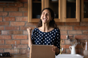 Emotional happy young hispanic woman unpacking huge cardboard box, feeling excited of getting...
