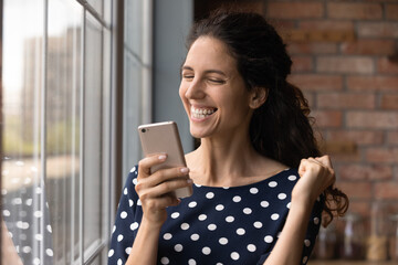 Emotional happy young latin woman making yes gesture, celebrating online lottery auction betting win looking at cellphone screen, feeling excited of internet success, standing near window at home.