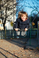Pięcioletni szczęśliwy chłopiec, blondyn z dłuższymi włosami huśtający się na huśtawce na osiedlowym placu zabaw. Zabawa na placu zabaw jesienią
