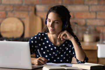 Happy interested young hispanic woman in wireless headphones looking at laptop screen, watching...