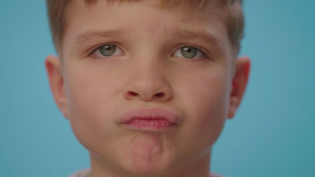 Close up of upset boy showing disappointment standing on blue background. Unhappy kid making lips grimaces.