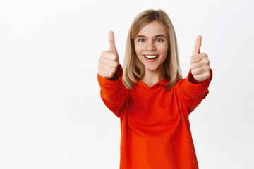 Cheerful blond little girl shows thumbs up in approval, smiling pleased and making compliment, satisfied by smth good, standing over white background