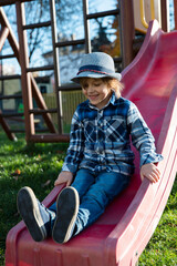Wesoły chłopiec w kapeluszu na czerwonej zjeżdżalni. Plac zabaw osiedlowy skąpany w jesiennym słońcu.