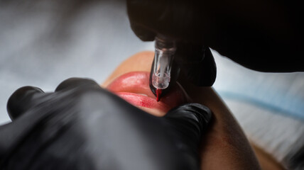 Cosmetologist in black gloves making permanent makeup on woman's lips with tattoo pen machine. Microblading process - obrazy, fototapety, plakaty
