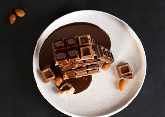 Melted liquid chocolate pours on a plate with pieces of chocolate bars with almond and hazelnut. Close-up.
