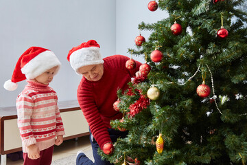 grandmother and granddaughter in Santa hats and red sweaters dress up a Christmas tree with Christmas toys
