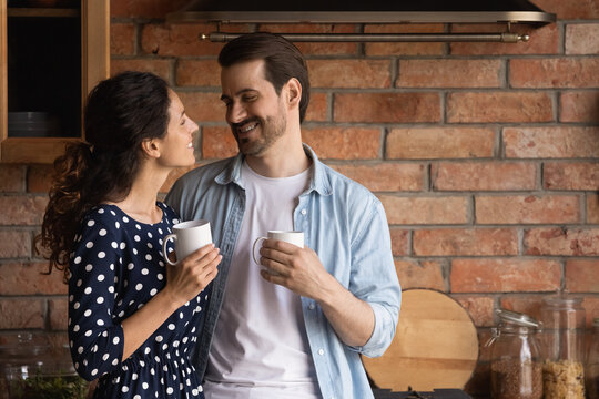 Romantic affectionate happy young family couple holding cup of tea or hot coffee, holding pleasant conversation in old-fashioned kitchen, enjoying carefree morning leisure time together at home.