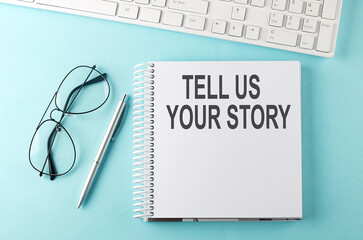Keyboard, notebook,pen and glasses on the blue background , text TELL US YOUR STORY
