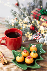 A red mug with black coffee, a plate in the shape of a Christmas tree with cupcakes and a Christmas decoration with illumination are on a wooden table. 