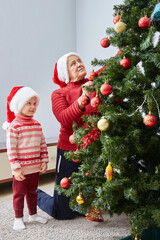 smiling granddaughter and grandmother in Santa hats and red sweaters dress up a Christmas tree with Christmas toys. Vertical frame.