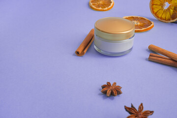 Fototapeta na wymiar Spa concept. Glass jar of cream, scrub with beige plastic lid, cinnamon, coriander, dry orange slice on purple background