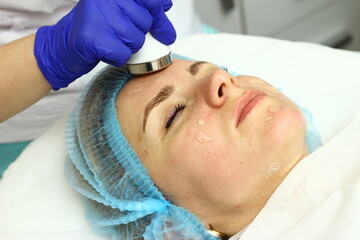 Beauty salon. A cosmetologist in medical gloves and protective mask doing a hydra peeling procedure on the client's cheeks. Side view. Close up. Professional skin care during coronavirus pandemic.