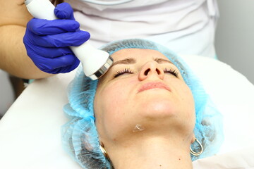 Naklejka premium Beauty salon. A cosmetologist in medical gloves and protective mask doing a hydra peeling procedure on the client's cheeks. Side view. Close up. Professional skin care during coronavirus pandemic.