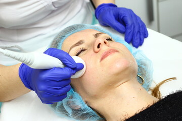 Fototapeta na wymiar Beauty salon. A cosmetologist in medical gloves and protective mask doing a hydra peeling procedure on the client's cheeks. Side view. Close up. Professional skin care during coronavirus pandemic.