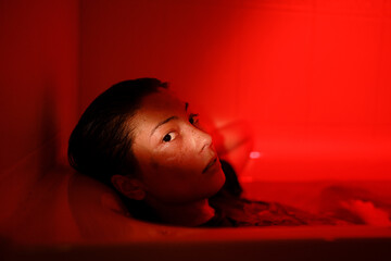 Portrait in red neon light of young sensual asian woman relaxing in bath at home, female looking at...