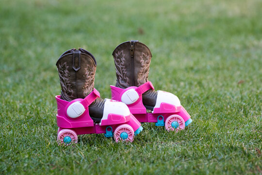 Empty Kids Cowgirl Cowboy Boots Shoes In A Pair Of Children's Pink Plastic Roller Skates On Green Grass. No People.