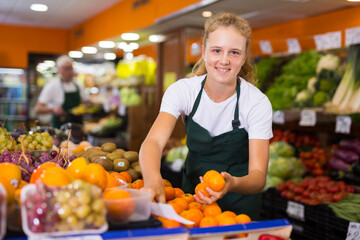 Young girl shopping assistant at her first job selling tangerines and other fruits in vegetable shop