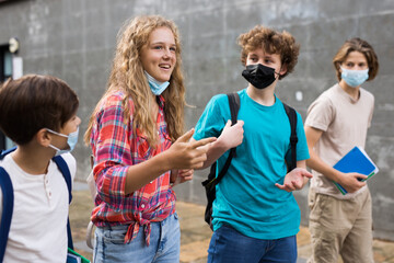 Group of teenage students in protective face masks discuss past lessons at a college on the street in summer day