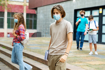 Teen boy in face mask with backpack going to home after finishing lessons at school on sunny day