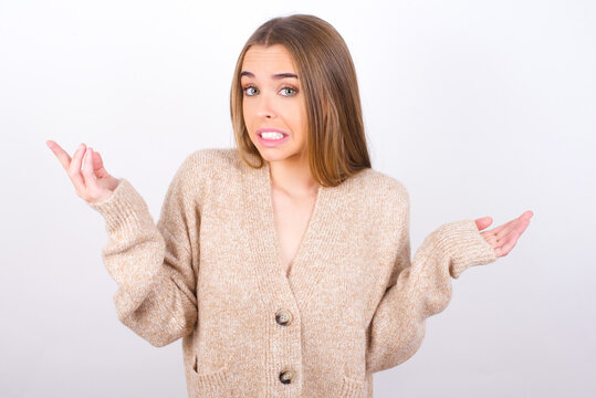 Clueless young caucasian girl wearing knitted sweater over white background shrugs shoulders with hesitation, faces doubtful situation, spreads palms, Hard decision