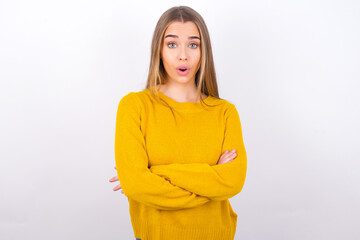 Shocked embarrassed Young caucasian girl wearing yellow sweater over white background keeps mouth widely opened. Hears unbelievable novelty stares in stupor