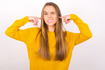 Happy Young caucasian girl wearing yellow sweater over white background ignores loud music and plugs ears with fingers asks to turn off sound