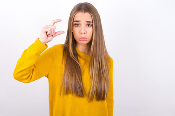 Young caucasian girl wearing yellow sweater over white background purses lip and gestures with hand, shows something very little.