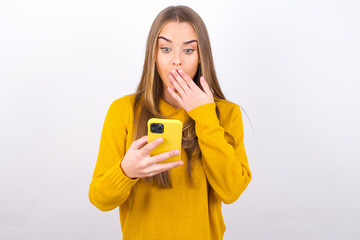 Young caucasian girl wearing yellow sweater over white background being deeply surprised, stares at...