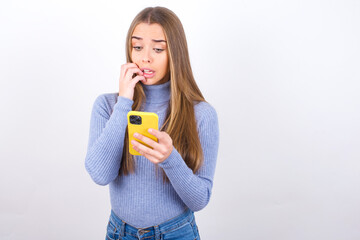 Portrait of pretty frightened Young caucasian girl wearing blue turtleneck over white background chatting biting nails after reading some scary news on her smartphone.