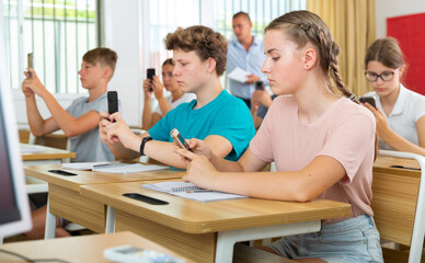 Young students with smartphones sitting in class room. They photographing something in front of them.