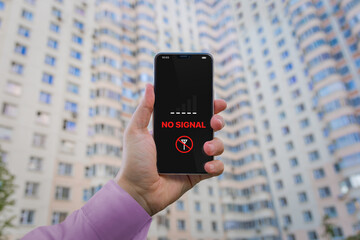 Message 'No signal' on the smartphone. A man's hand holds a smartphone against the background of a residential high-rise building. Problems with cellular coverage in a densely populated area