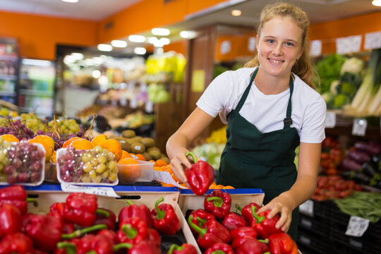 Fifteen-year-old girl who works part-time in a store as a trainee saleswoman puts red bell pepper on the counter for sale