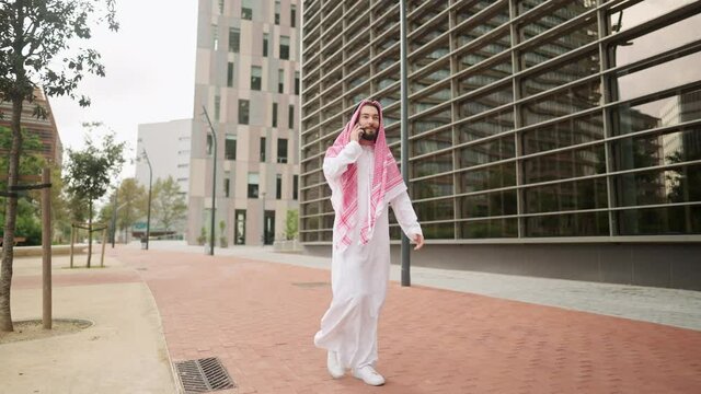 Saudi man with smartphone outdoors, arab businessman talking on mobile phone while walking on road
