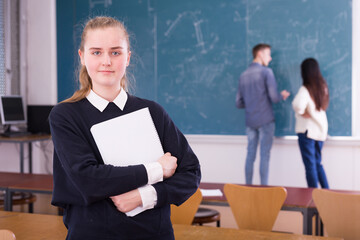 Portrait of girl student standing with notebook in classroom, students writing on whiteboard
