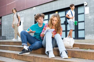 Positive schoolchildren preparing schoolwork, sitting on steps outside school in sunny autumn day