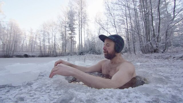 An ice bather enters the ice hole and sits down up to his neck, breathing